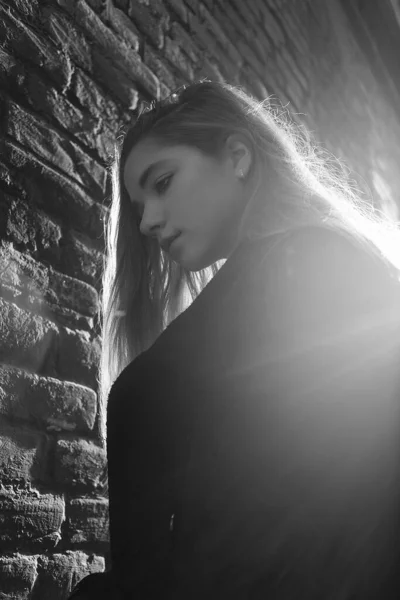 Νεαρό κορίτσι με μαύρο παλτό στέκεται μπροστά από έναν τοίχο από τούβλα στο όμορφο φως του ηλιοβασιλέματος. Έννοια της μοναξιάς — Φωτογραφία Αρχείου