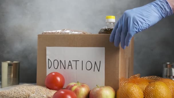 Voluntario usando guantes protectores poniendo comida de comestibles en caja de donación de cartón — Vídeos de Stock