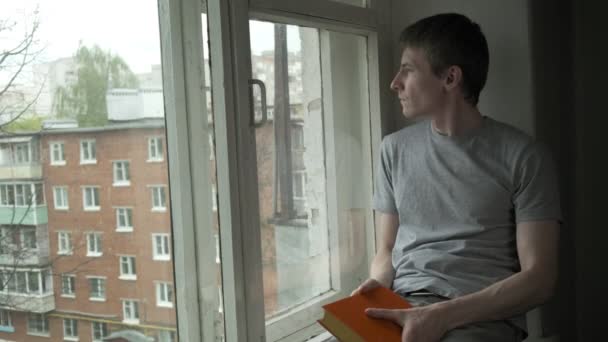 Joven hombre aburrido sentado en el alféizar de la ventana, mirando a la ventana, sosteniendo el libro en las manos — Vídeos de Stock
