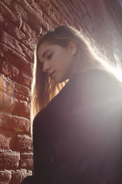 Молодая девушка в черном пальто стоит перед кирпичной стеной в красивом подсветке заката. Концепция одиночества — стоковое фото