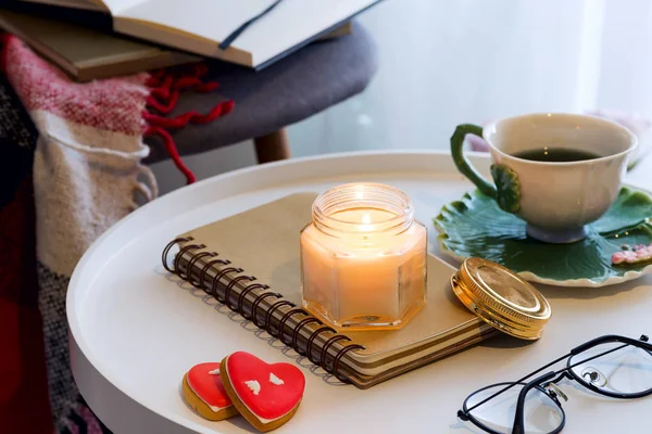 Una vela con un cuaderno, una taza de té en una mesa con una silla, una manta a cuadros y un libro. Interior del hogar — Foto de Stock