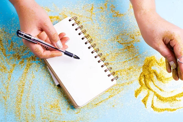 Mujeres doradas manos brillantes están tomando brillo agold y escribe en un cuaderno. Fondo festivo azul con brillo . — Foto de Stock