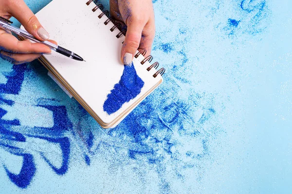 Las manos brillantes azules de las mujeres están tomando brillo agold y escribe en un cuaderno. Fondo festivo azul con brillo . — Foto de Stock