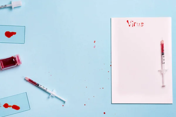 Jeringas y probeta con sangre sobre fondo azul con papel blanco y la inscripción Virus — Foto de Stock