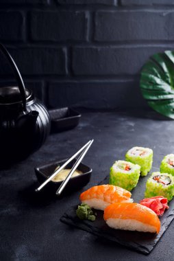 Taze hazırlanmış suşi ve rulo seti siyah bir tahta üzerinde servis edilmiş metal yemek çubukları, koyu beton bir arka planda siyah seramik çay demliği yeşil yapraklı, kopyalama alanı. Japon yemeği..