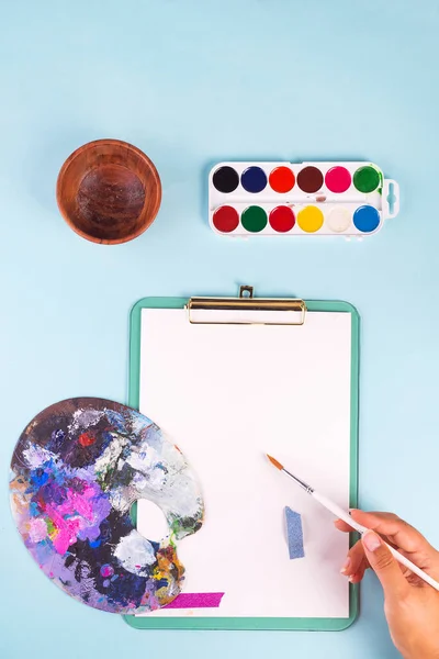 Womans hand with paintbrush above blank white paper sheet on a light blue background. Flat lay. — 스톡 사진