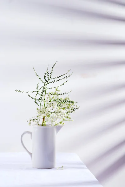 Galhos de flores frescas de planta de Gypsophila em vaso de vidro em uma mesa coberta de tecido contra parede com sombras de jalousie . — Fotografia de Stock