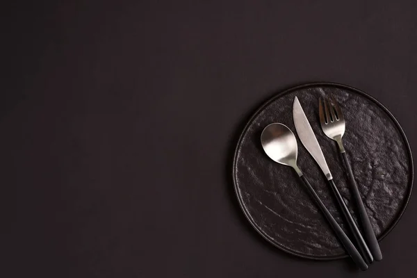 Schwarze handgefertigte Keramikteller serviert Gabel, Löffel und Messer auf dem gleichen Farbhintergrund. — Stockfoto