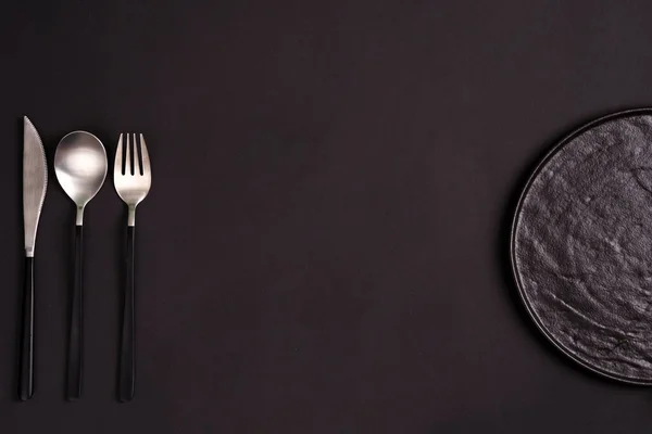 Black handmade ceramic plate served fork, spoon and knife on the same color background. — Stock Photo, Image