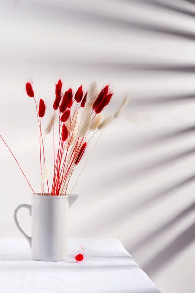 Caneca de cerâmica com flores secas macias de outono vermelho e branco grama Lagurus Ovatus em uma mesa coberta de tecido contra parede com sombras de jalousie . — Fotografia de Stock