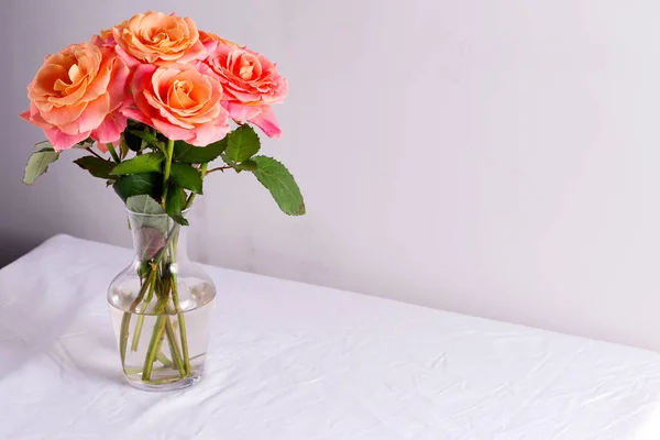 Üvegváza gyönyörű csokor friss természetes rózsa virágok az asztalon borított fehér ruha ellen világosszürke fal. — Stock Fotó