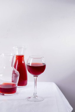 Şarap kadehinde kırmızı şarap, beyaz tekstil arka planında gölgeli sürahi ve şişe.