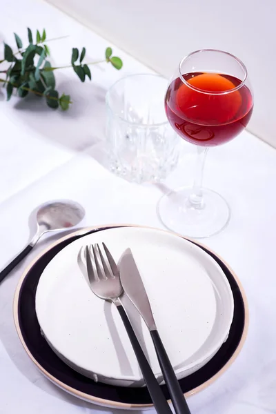 Geschirr und Dekorationen zum Servieren einer festlichen Tafel. Teller, Rotweinglas und Besteck mit grünen Blättern auf weißem Textilhintergrund. — Stockfoto