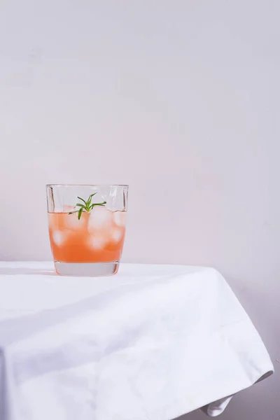 Cóctel rosa con romero y hielo en vaso sobre un mantel blanco sobre la mesa —  Fotos de Stock