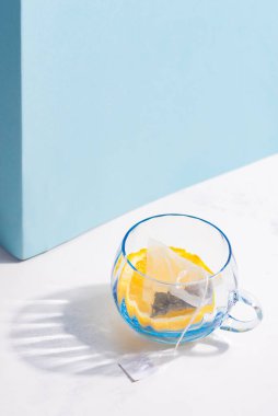 Bardakta meyve çayı poşeti ve beyaz bir masada limon dilimi ve pastel mavi duvarda gölgesi var..