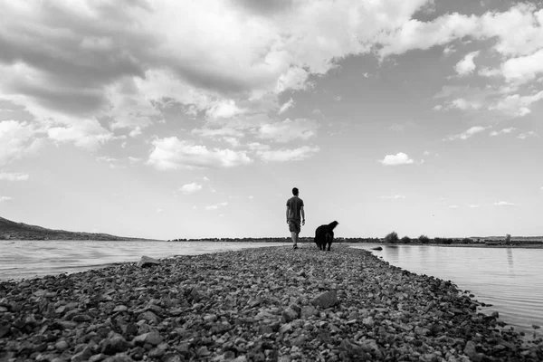 Man and his dog walking on the sandy path across the lake on a b — Stock Photo, Image