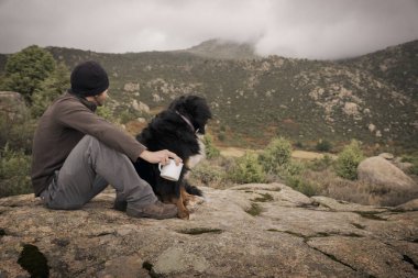 Genç adam onun köpek ve bir kupa ile dağ başında oturur