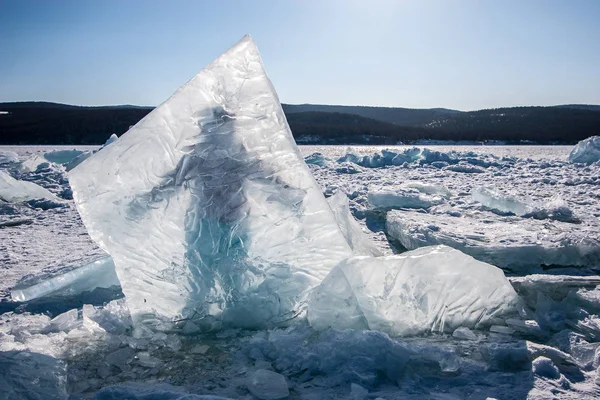 A huge ice piece frozen into Lake Baikal, behind which a man stands. Clear skies, hills behind a lake with snow and scattered ice.