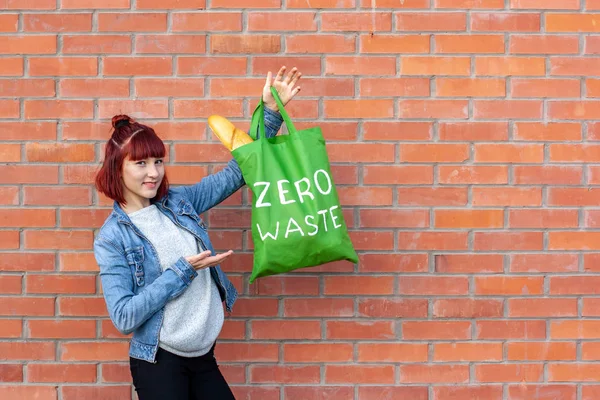 A young girl shows a textile eco bag zero waste on a brick wall background. The girl smiles, red short hair. French bread in a bag. Copy space on the right.