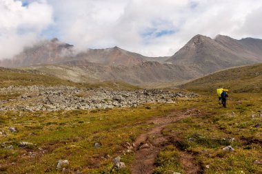 Büyük sırt çantalı turistler dağlarda yürüyüş parkuruna giderler. Yüksek dağların tepesindeki bulutlar. Taş talus. Yokuşta yeşil çimenler. Yatay.