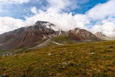 Bulutların arasında taşlarla kaplı büyük bir dağ. Bulutlu mavi gökyüzü. Yamacında taşlar olan otlar. Yatay.