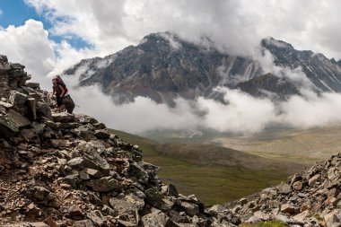 Kayalık bir yamaç, büyük bir sırt çantası ve çubuğu olan bir turist dağa tırmanıyor. Bulutlar uzakta büyük bir dağı gizler. Aşağıda yeşil bir vadi var. Yatay.
