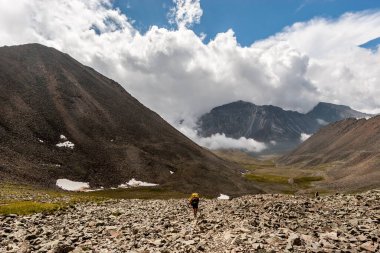 Sırt çantalı bir yürüyüşçü yerdeki yüksek dağların arasında yürüyor. Yüksek dağlar ve alçak beyaz bulutlar. Dağların arasındaki vadi. Yatay.