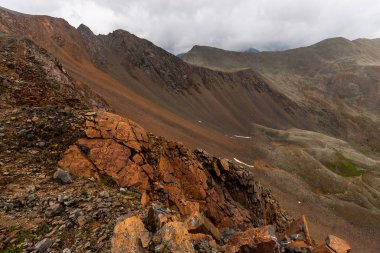 Dik bir uçurumdan ve tepesinde bulutlar olan bir sıradağdan manzara. Ön planda kırmızı çatlamış taşlar var. Yatay.
