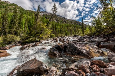 Büyük taşlar ve aralarında akan bir dağ nehri. Ormanın ve dağların etrafında. Mavi gökyüzü ve dağların üzerinde alçak beyaz bulutlar. Yatay.