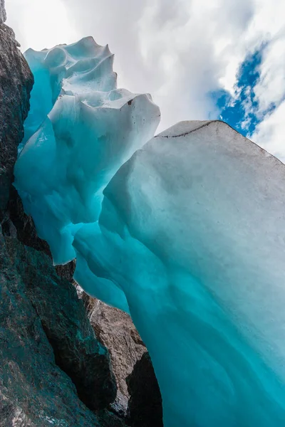 Beautiful ice of blue color hanging from stones. A small stream of water is visible under the ice. Blue sky with clouds. Vertical.