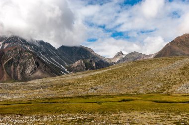 Dağ manzarası. Tepelerinde bulutlar olan bir sıra yüksek uçurum. Yerde bir sürü taş ve yeşil çimen var. Mavi gökyüzü. Yatay.