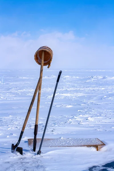 Tools for punching an ice hole in ice. An ice pick, a shovel and a bucket are stuck in the ice hole against a snowy plain and blue sky. Vertical.