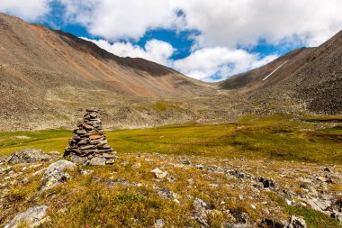 Önünde büyük bir cairn bulunan dağ manzarası. Dağların üzerinde beyaz bulutlar olan mavi gökyüzü. Dağlardaki bulutların gölgesi. Yatay.