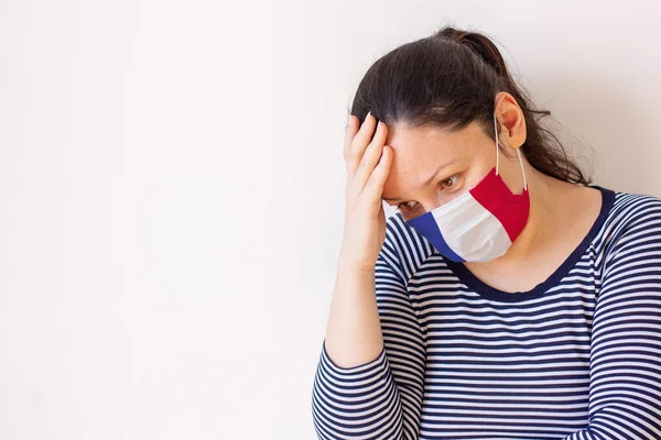 The girl in the medical mask painted like the flag of France thoughtfully lowered her head and put her hand to her head. Looks down to the side. Copy space on a white background. Horizontal.