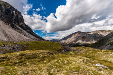Yamaçta yemyeşil bitki örtüsü ve beyaz yosun var. Uzakta yüksek dağlar ve mavi gökyüzünde alçak beyaz bulutlar. Yatay.