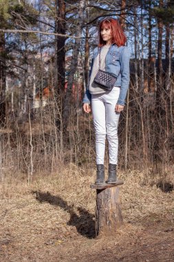 Genç kızıl saçlı bir kız ormanda bir kütüğün üzerinde duruyor. Avrupa görünüşü. Kot ceket ve beyaz pantolon. Çantayı omzuna koy. Dikey.
