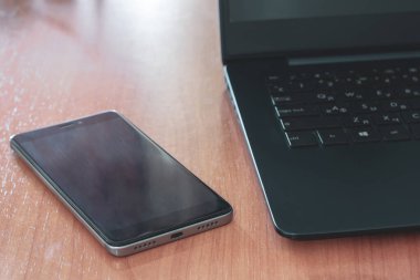 Masada siyah dizüstü bilgisayar ve cep telefonu var. Seçici sığ bir şekilde telefona odaklan. Arka ışık. Yatay.