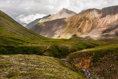 Dağların arasındaki yeşil vadide bir yarık var. Etrafta güzel dağlar var. Yere, yosun ve çimenler. Hava bulutlu. Yatay.
