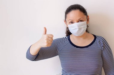 Tıbbi maskeli bir kız, arka planda fotokopi alanı olan beyaz bir başparmak gösteriyor. Avrupa görünüşü. Uzun siyah saçlı. Çizgili ceket. Yüze odaklan. Yatay.