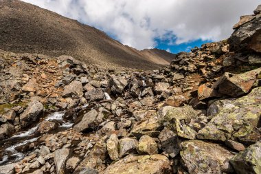 Dağlardaki büyük taşların arasından akan küçük bir nehir. Dağların arasında. Mavi gökyüzü ve bulutlar. Yatay.
