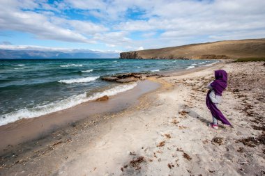 Ekoseye sarılı bir kız Baykal Gölü 'nün kumlu kıyısında duruyor. Rüzgârlı ve dalgalı. Ufuktaki dağlar. Mavi gökyüzünde bulutlar. Yatay.