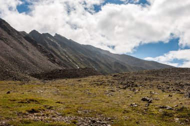 Dağ sıraları ve dağ yamacında taşlar ve yeşil otlar. Mavi gökyüzü ve bulutlar. Yatay.