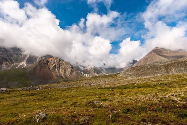 Bulutlarda yüksek tepeler olan tepe tarafında. Taştan nehirleri olan kayalar. Mavi gökyüzü. Yokuşta yeşil çimenler. Bir sürü taş. Yatay.