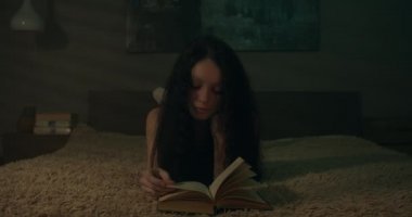 Loş bir odada kitap okuyan genç bir kadın. Bir sayfa çeviriliyor.