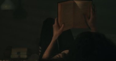 Loş bir odada kitap okuyan genç bir kadın. Omzundan vurulmuş. El bilgisayarı.