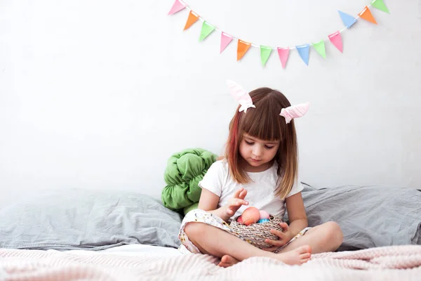 Cute little girl wearing bunny ears holding painted Easter eggs and smiling at home interior. Happy easter, spring equinox concept. — Stock Photo, Image