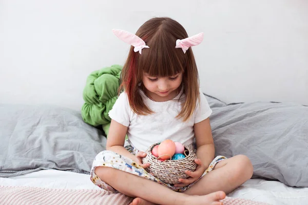 Χαριτωμένο κοριτσάκι φορώντας αυτιά λαγουδάκι κρατώντας βαμμένα αυγά του Πάσχα και χαμογελώντας στο εσωτερικό του σπιτιού. Χαρούμενο Πάσχα, έννοια της εαρινής ισημερίας. — Φωτογραφία Αρχείου