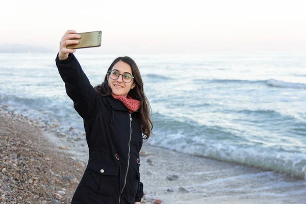 Młoda kaukaska dziewczyna z okulary robi selfie na plaży w zimie — Zdjęcie stockowe
