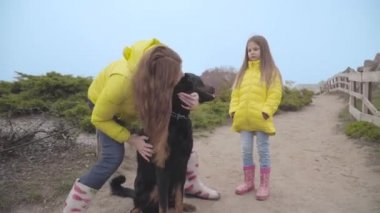Sarı paltolu genç beyaz kadın sonbahar parkında büyük siyah dobermanı okşuyor ve yanında sarı ceketli küçük kız duruyor. Aile hafta sonlarını sadık bir evcil hayvanla dışarıda geçiriyor.