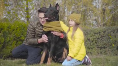 Beyaz bir erkek ve küçük bir kızın portresi. Yeşil çimlerde oturuyor. Hardal atkısı içinde zarif bir doberman. Erkek ve kız kardeşler köpekleriyle dışarıda vakit geçiriyorlar.